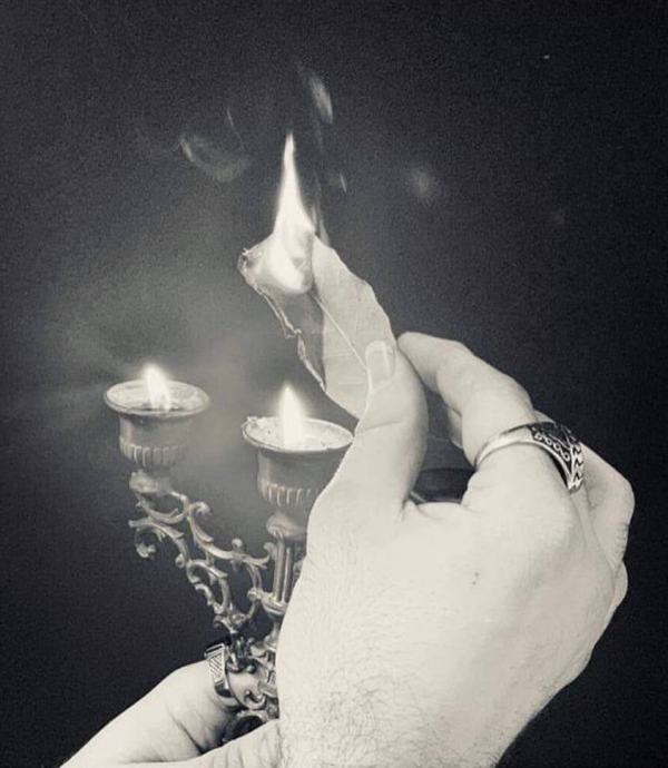 Candelabro retto da una mano con sopra di esso una foglia di salvia che brucia grazie alle fuoco delle candele, sorretta a sua volta dalla mano destra. Foto in bianco e nero.